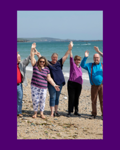 A group of volunteers with people with a learning disability on a beach with their hands raised and smiling